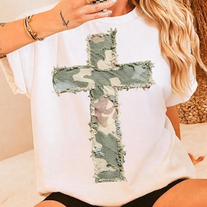 May include: White short-sleeved t-shirt featuring a large cross design. The cross is made of camouflage fabric with frayed edges, creating a textured appearance. The shirt is a casual, everyday wear item.