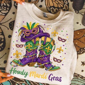 Könnte beinhalten: Ein hellbeige Sweatshirt mit einem farbenfrohen Mardi Gras-Design. Das Design umfasst lila Cowboy-Stiefel, einen passenden Hut und eine Maske, die alle mit Perlen und Fleur-de-Lis-Symbolen verziert sind. Die Worte "Howdy Mardi Gras" sind in Grün und Gelb geschrieben.