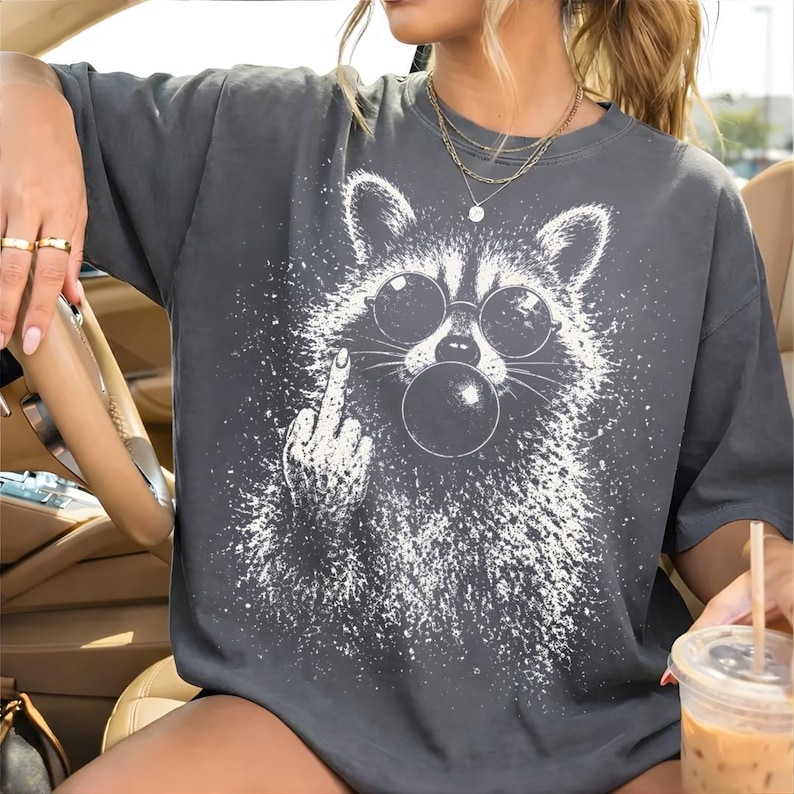 Pu&ograve; includere: T-shirt grigio scuro con una grafica bianca di un procione con occhiali da sole che fa una bolla. Il procione fa il dito medio. La maglietta ha una vestibilit&agrave; ampia e maniche corte.