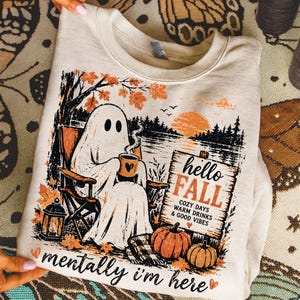 Op de afbeelding: Een crèmekleurig sweatshirt met een afbeelding van een geest die in een stoel bij een meer zit en een mok vasthoudt. Het ontwerp bevat oranje herfstbladeren, pompoenen, een lantaarn en een houten bord met de tekst hello FALL COZY DAYS WARM DRINKS & GOOD VIBES. De tekst eronder zegt mentally i'm here.