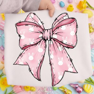 May include: A large pink bow with a white bunny pattern. The bow is held up by a person's hand. The bow is a light pink color with a darker pink outline. The bunny pattern is white. The bow is a decorative item.