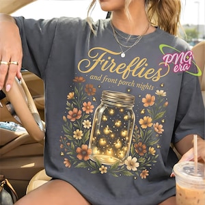 Può includere: T-shirt grigio scuro con le parole "Fireflies and front porch nights" in oro. La maglietta presenta un barattolo di vetro pieno di lucciole luminose, circondato da fiori arancioni, gialli e bianchi.