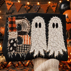 May include: A black sweatshirt featuring a large applique design that spells out the word BOO. The letter B is constructed from various fabric patches including plaid, spiderweb, and ghost patterns. Two ghosts made of white lace are featured.