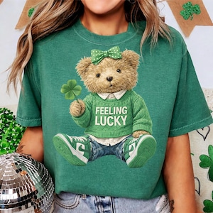 May include: Green t-shirt featuring a teddy bear wearing a green sweater with the words "Feeling Lucky". The bear holds a four-leaf clover and wears a green bow and green sneakers. The shirt is a St. Patrick's Day theme.