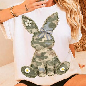 Pode incluir: Camiseta branca com um design de coelho camuflado, um laço xadrez azul e detalhes de margaridas. O coelho é feito de tecido com um padrão verde, marrom e azul. Uma peça casual para o dia a dia.