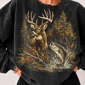 May include: A charcoal grey acid-wash crewneck sweatshirt featuring a detailed graphic print of a large buck with antlers standing in a forest, alongside a fish jumping out of the water amidst reeds and tall grasses in shades of gold and brown.