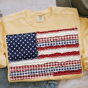 May include: A yellow sweatshirt with a patchwork American flag design. The flag features a blue square with white stars and red, white, and blue striped sections. The sweatshirt has a crew neck and long sleeves.