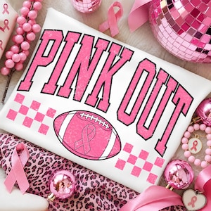 May include: A white t-shirt featuring the text PINK OUT in distressed pink letters with black outlines. Below the text is a pink football graphic with a pink awareness ribbon in the centre, flanked by pink checkered patterns on both sides.