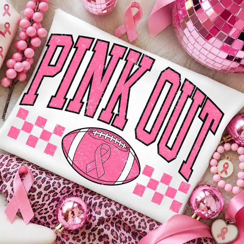 May include: A white t-shirt featuring the text PINK OUT in distressed pink letters with black outlines. Below the text is a pink football graphic with a pink awareness ribbon in the centre, flanked by pink checkered patterns on both sides.