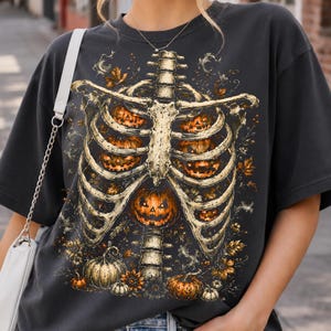 May include: A charcoal gray short-sleeve t-shirt featuring a graphic print of a human rib cage with glowing orange jack-o'-lanterns nestled between the bones, surrounded by autumn leaves, sunflowers, and small pumpkins at the base.
