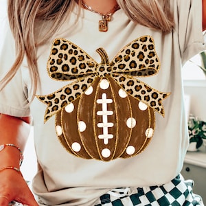May include: A beige t-shirt featuring a graphic of a brown pumpkin with white polka dots and a football lace design in the center. The pumpkin is topped with a large leopard print bow. The shirt has short sleeves and a crew neckline.