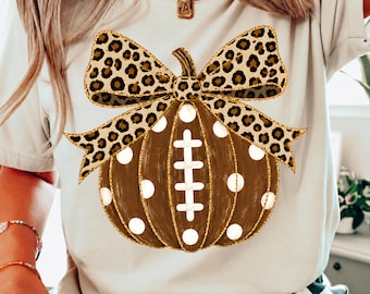 May include: A beige t-shirt featuring a graphic of a brown pumpkin with white polka dots and a football lace design in the center. The pumpkin is topped with a large leopard print bow. The shirt has short sleeves and a crew neckline.