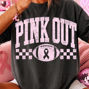 May include: A charcoal grey short-sleeve t-shirt featuring the text PINK OUT in a distressed light pink font. Below the text is a graphic of a football with a black awareness ribbon in the centre, flanked by two light pink checkered patterns.
