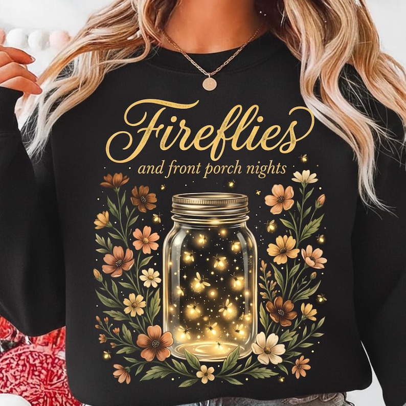 Pu&ograve; includere: Felpa nera con un disegno di un barattolo pieno di lucciole luminose, circondato da fiori marroni, arancioni e crema. Il testo "Fireflies and front porch nights" &egrave; scritto in oro.