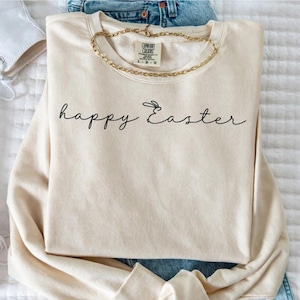 May include: A cream-colored sweatshirt with the words "happy Easter" in black script. A gold chain necklace rests on the sweatshirt. The sweatshirt is folded on top of blue jeans.