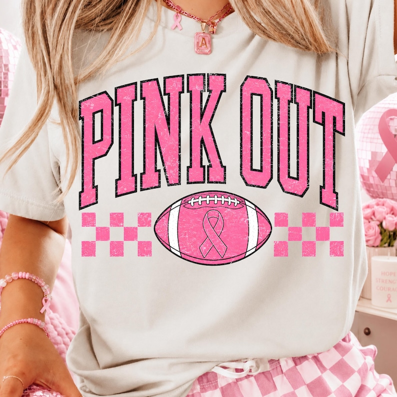 May include: A light-coloured t-shirt featuring the text PINK OUT in large, distressed pink letters. Below the text is a pink football graphic with a pink awareness ribbon in the centre, flanked by pink and white checkered patterns on both sides.