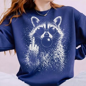Pu&ograve; includere: Felpa blu navy con grafica bianca di un procione con occhiali da sole e che soffia una bolla. Il procione fa il dito medio. Il design ha un effetto invecchiato e macchiato.