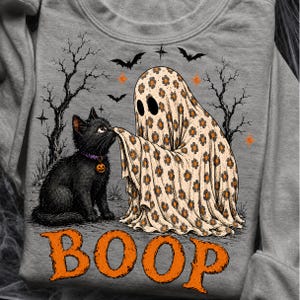 May include: A heather gray sweatshirt featuring a graphic of a black cat with a purple collar and a ghost covered in a beige sheet with an orange and black flower pattern. The ghost touches the cat's nose with the text BOOP in orange.