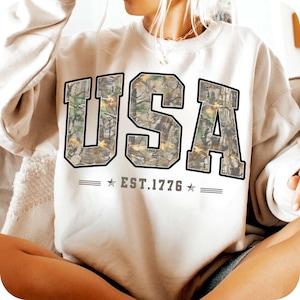 May include: Cream-colored sweatshirt with the word "USA" in large, block letters. The letters are filled with a camouflage pattern and outlined in black. Below the letters is the text "EST. 1776" with stars on either side.