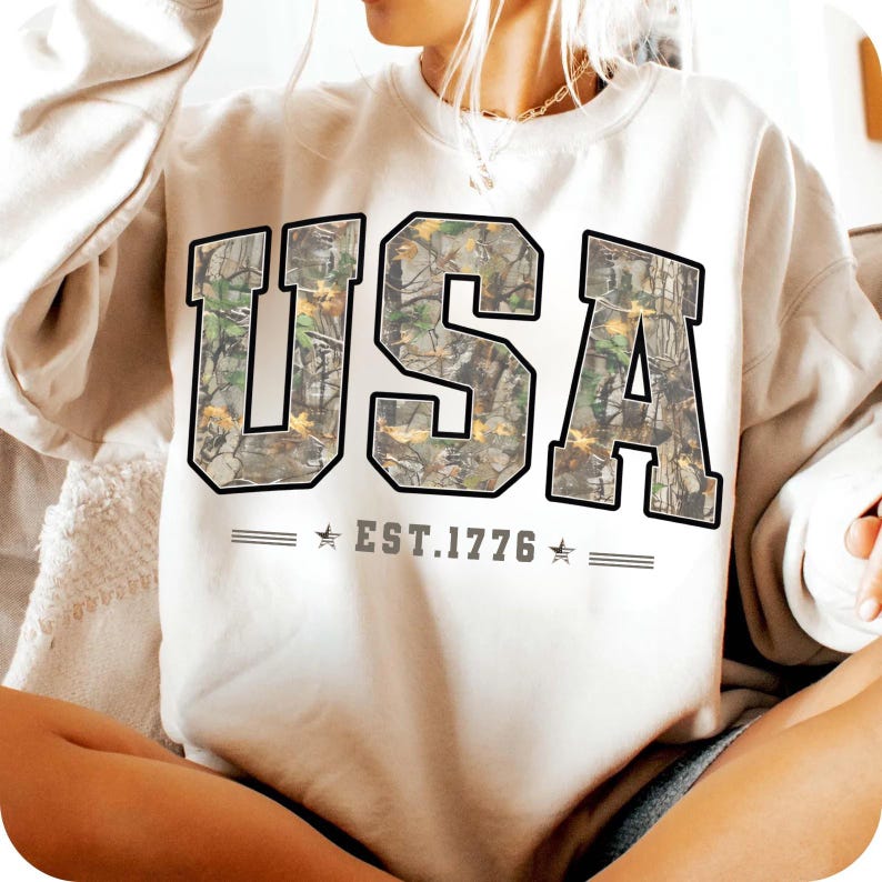 May include: Cream-colored sweatshirt with the word "USA" in large, block letters. The letters are filled with a camouflage pattern and outlined in black. Below the letters is the text "EST. 1776" with stars on either side.