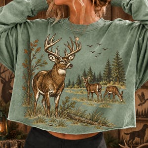 May include: A sage green long-sleeve sweatshirt featuring a graphic print of a large buck with antlers standing in a forest clearing, accompanied by two smaller deer, tall pine trees, and a flock of birds flying in the sky above the landscape.