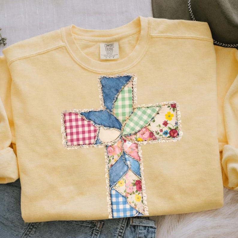 May include: A pale yellow sweatshirt featuring a patchwork cross. The cross is constructed from diverse fabrics, including denim, gingham, and floral patterns. The sweatshirt has a crew neck and long sleeves.