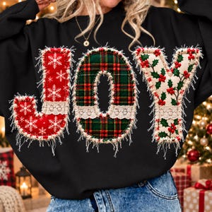 May include: A black long-sleeve sweatshirt featuring the word JOY in large, appliqued letters. The J is red with white snowflakes, the O is green and red plaid, and the Y is white with a red holly berry and green leaf pattern.