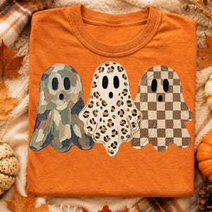 May include: An orange crew neck t-shirt featuring a graphic of three ghosts. The left ghost has a green and brown camouflage pattern, the middle ghost has a tan and black leopard print, and the right ghost has a brown and tan checkerboard pattern.