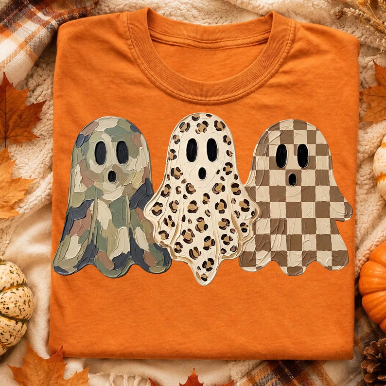 May include: An orange crew neck t-shirt featuring a graphic of three ghosts. The left ghost has a green and brown camouflage pattern, the middle ghost has a tan and black leopard print, and the right ghost has a brown and tan checkerboard pattern.