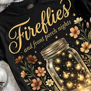 Pu&ograve; includere: T-shirt nera con un design grafico raffigurante un barattolo pieno di lucciole luminose. Le parole "Fireflies and front porch nights" sono scritte in oro sopra il barattolo, circondate da fiori.