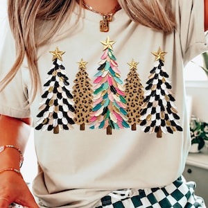 May include: A beige short-sleeve t-shirt featuring a graphic print of five stylized Christmas trees. The trees are decorated with gold stars at the top and display a mix of black, white, gold, pink, blue, and leopard print patterns.