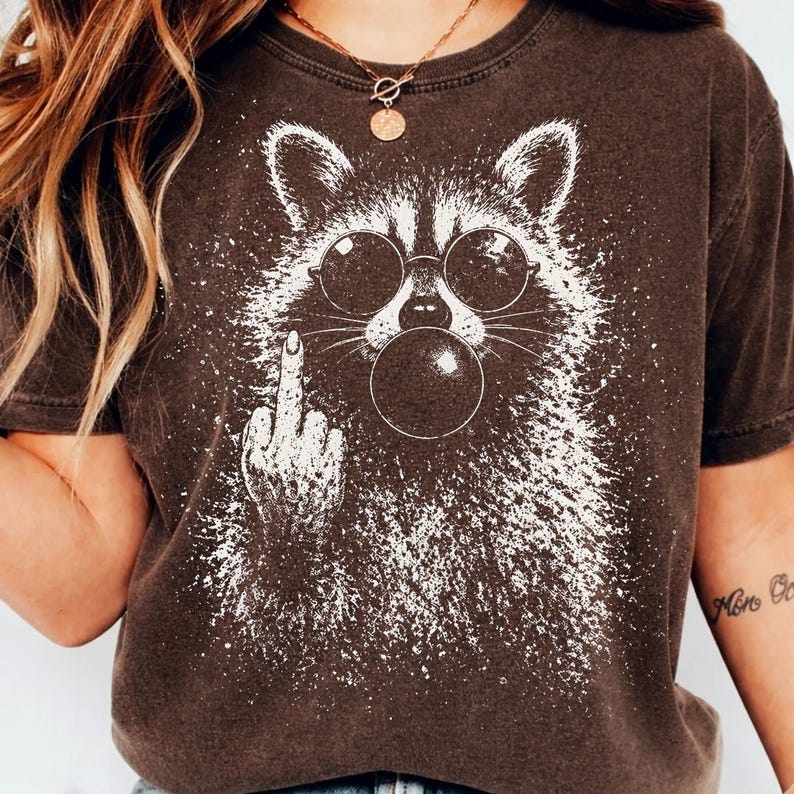 Pu&ograve; includere: T-shirt marrone con grafica bianca di un procione con occhiali da sole che fa una bolla. Il procione fa il dito medio. La maglietta ha un aspetto vintage e usurato.