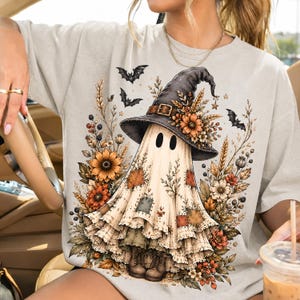 May include: A light beige t-shirt featuring a graphic of a ghost wearing a black witch hat with a buckle, surrounded by autumn flowers, wheat stalks, and small black bats. The ghost has patches on its fabric body and wears brown boots.