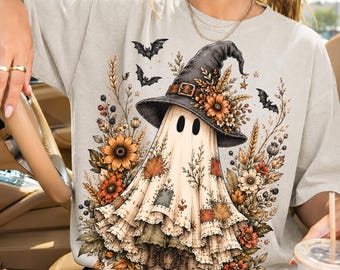 May include: A light beige t-shirt featuring a graphic of a ghost wearing a black witch hat with a buckle, surrounded by autumn flowers, wheat stalks, and small black bats. The ghost has patches on its fabric body and wears brown boots.