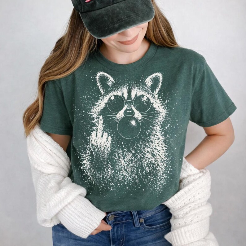 Pu&ograve; includere: T-shirt verde scuro con grafica bianca di un procione con occhiali da sole che soffia una bolla. Il procione fa il dito medio. La maglietta &egrave; a girocollo.