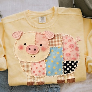 May include: A light yellow sweatshirt featuring a patchwork pig design. The pig has a beige face, pink ears and cheeks, and a pink and brown patterned body. The sweatshirt has a crew neck and the Comfort Colors brand tag.