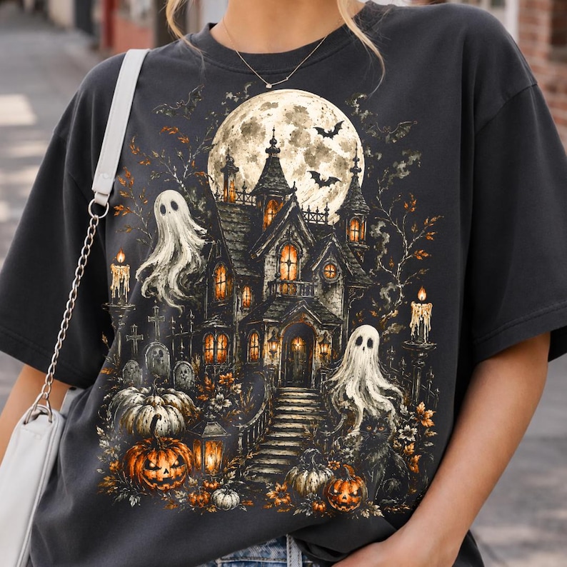 May include: A charcoal gray t-shirt featuring a detailed graphic print of a haunted mansion under a full moon. The design includes two white ghosts, orange pumpkins, lit candles, and flying bats, creating a dark, atmospheric Halloween scene.