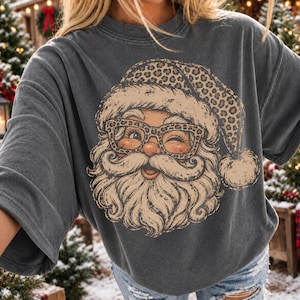 Può includere: Una t-shirt a maniche corte color grigio antracite con una stampa grafica di Babbo Natale che indossa un cappello leopardato e occhiali abbinati. L'illustrazione presenta una barba dettagliata, baffi e un'espressione ammiccante.
