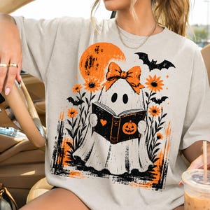 Pu&ograve; includere: Una t-shirt beige con la grafica di un fantasma bianco che indossa un fiocco arancione e legge un libro nero con l'icona di una zucca. Il design include una luna arancione, pipistrelli neri e girasoli arancioni in uno stile artistico.