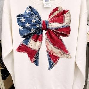 K&ouml;nnte beinhalten: Wei&szlig;es Sweatshirt mit einem gro&szlig;en Schleifendesign. Die Schleife besteht aus Denim und rot-wei&szlig;em Stoff, der der amerikanischen Flagge &auml;hnelt. Das Design umfasst wei&szlig;e Sterne auf blauem Denim-Hintergrund und rot-wei&szlig;e Streifen.