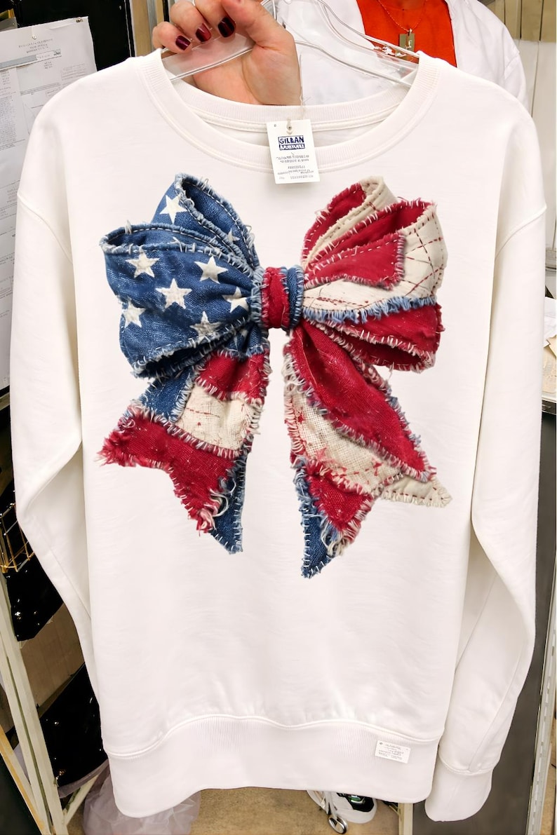 K&ouml;nnte beinhalten: Wei&szlig;es Sweatshirt mit einem gro&szlig;en Schleifendesign. Die Schleife besteht aus Denim und rot-wei&szlig;em Stoff, der der amerikanischen Flagge &auml;hnelt. Das Design umfasst wei&szlig;e Sterne auf blauem Denim-Hintergrund und rot-wei&szlig;e Streifen.