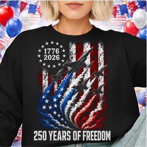 Pu&ograve; includere: Felpa nera con un design della bandiera americana, jet e il testo "1776 2026" e "250 YEARS OF FREEDOM". Il design della bandiera utilizza rosso, bianco e blu, con stelle e strisce. I jet lasciano scie di fumo rosso, bianco e blu.