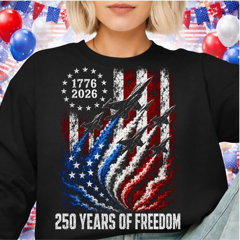 Pu&ograve; includere: Felpa nera con un design della bandiera americana, jet e il testo "1776 2026" e "250 YEARS OF FREEDOM". Il design della bandiera utilizza rosso, bianco e blu, con stelle e strisce. I jet lasciano scie di fumo rosso, bianco e blu.