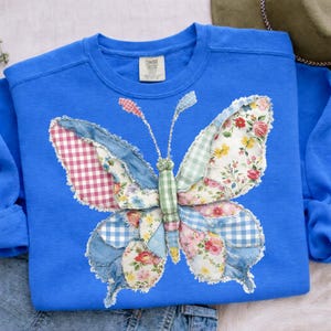 Könnte beinhalten: Blaues Sweatshirt mit einem Patchwork-Schmetterlingsdesign. Die Schmetterlingsflügel zeigen eine Mischung aus floralen, Vichy- und Denim-Stoffen. Das Sweatshirt hat einen Rundhalsausschnitt und lange Ärmel. Das Etikett lautet "Comfort Colors".
