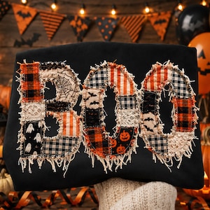 May include: A black sweatshirt featuring the word BOO appliqued in a patchwork style. The letters are constructed from various fabrics including orange and black plaid, white lace, and black prints with ghosts, bats, spiderwebs, and pumpkins.