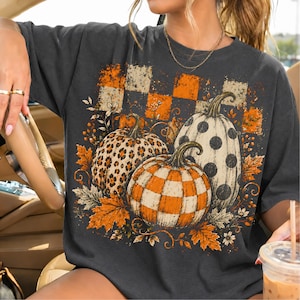 May include: A charcoal gray t-shirt featuring a graphic print of three pumpkins with different patterns: one with leopard print, one with orange and white checks, and one with black polka dots, surrounded by orange autumn leaves and flowers.
