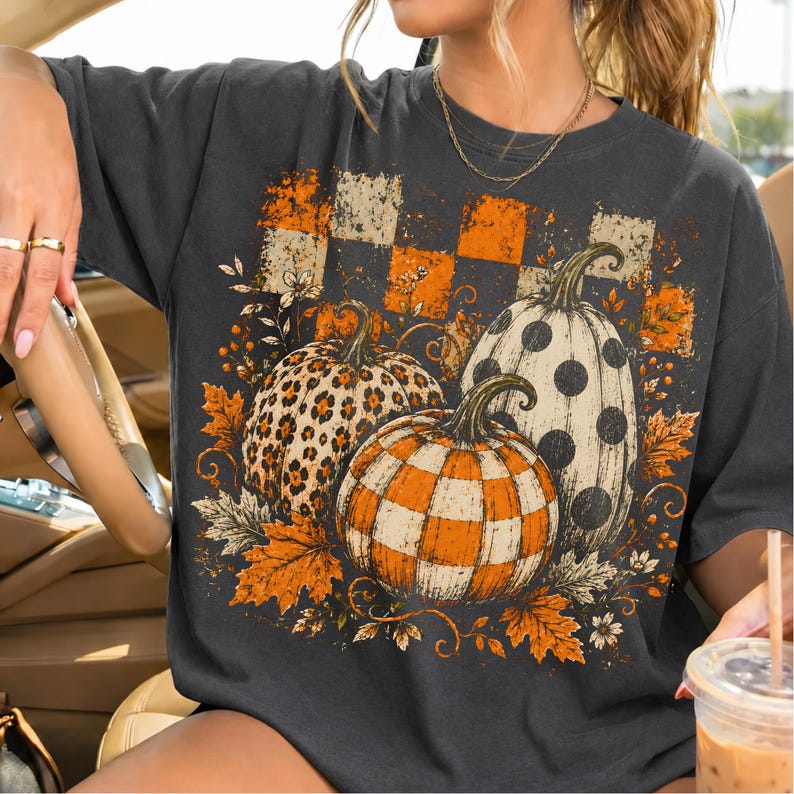 May include: A charcoal gray t-shirt featuring a graphic print of three pumpkins with different patterns: one with leopard print, one with orange and white checks, and one with black polka dots, surrounded by orange autumn leaves and flowers.