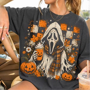 May include: A charcoal gray t-shirt featuring a graphic print of a hooded ghost figure wearing a plaid scarf and holding a pumpkin-faced coffee cup. The design includes orange and white checkered patterns, autumn maple leaves, and two jack-o'-lanterns.