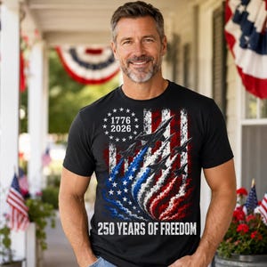 Pu&ograve; includere: T-shirt nero con un design patriottico raffigurante la bandiera americana, jet e il testo "1776 2026" e "250 YEARS OF FREEDOM". Il design incorpora i colori rosso, bianco e blu.