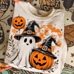 May include: A beige crewneck sweatshirt featuring a graphic print of a white ghost wearing a black witch hat and a jack-o'-lantern wearing a black witch hat. The design includes an orange pumpkin balloon, a flying bat, and orange checkered squares.